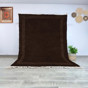 Op de afbeelding: Een groot, rechthoekig bruin tapijt met een donkerdere bruine rand en franjes langs de onderkant. Het tapijt wordt gepresenteerd tegen een witte muur, met een ingelijste foto en decoratieve wandkleden op de achtergrond.