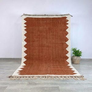 May include: A rectangular rug with a terracotta-colored center and a cream-colored border with a zigzag design. The rug has a fringe along the bottom edge and is displayed on a light gray floor.