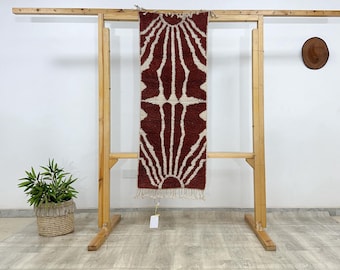 May include: A rectangular, hand-woven rug with a geometric design of white sunburst patterns on a deep red background. The rug is displayed on a wooden rack, showcasing its texture and design. The rug has a fringe at the bottom.