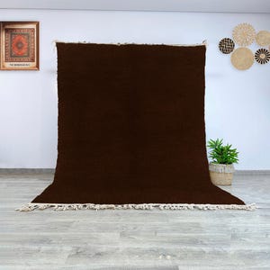 May include: A large, dark brown rug with a textured surface and a cream-colored fringe along the bottom edge. The rug is displayed against a white wall, with a framed artwork and decorative wall hangings visible in the background.