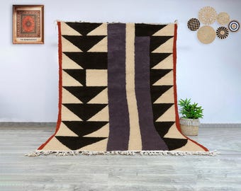 Könnte beinhalten: Ein großer, rechteckiger Teppich mit geometrischem Design. Der Teppich hat schwarze Dreiecke, einen zentralen lila Streifen und einen cremefarbenen Hintergrund. Die Ränder sind mit einem roten Rand und Fransen versehen.