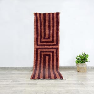 Op de afbeelding: Een lang, smal tapijt met een geometrisch patroon van bruine en roze strepen. Het tapijt heeft aan beide uiteinden franjes.