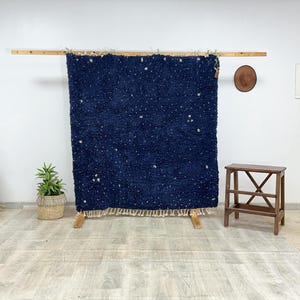 以下が含まれることがあります： 木製のフレームに展示された、斑点模様の濃い青色のラグ。 ラグは毛むくじゃらの質感で、下端にはクリーム色のフリンジが付いています。 小さな木製のスツールと鉢植えの植物が前景にあります。