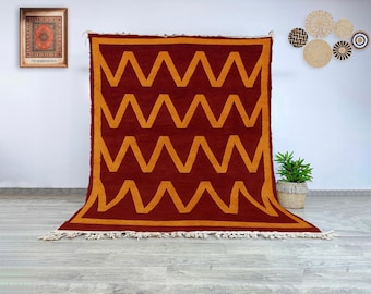 Könnte beinhalten: Ein großer, rechteckiger Teppich mit einem kräftigen geometrischen Design. Der Teppich hat einen tiefroten Hintergrund mit einem sich wiederholenden Muster aus goldgelben Zickzacklinien. Die Kanten sind mit Fransen versehen.