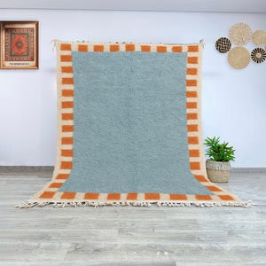 Könnte beinhalten: Ein großer, rechteckiger Teppich mit hellblauem Zentrum und einem orangefarbenen und cremefarbenen Rand. Der Teppich hat eine strukturierte Oberfläche und einen Fransenrand. Der Teppich wird vor einer weißen Wand ausgestellt.