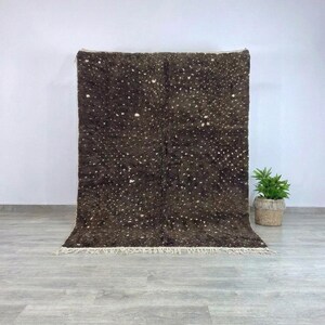 May include: A dark brown rug with a speckled pattern of white dots. The rug has a thick pile and a fringe along the bottom edge. The rug is displayed against a white wall and a light gray floor. A small potted plant is to the right.