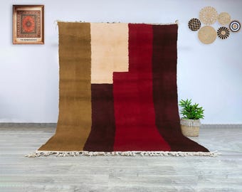Könnte beinhalten: Ein großer, rechteckiger Teppich mit geometrischem Design. Der Teppich hat vertikale Streifen in Braun-, Beige- und Rottönen. Der Teppich hat einen Fransenbesatz am unteren Rand. Der Teppich wird vor einer weißen Wand präsentiert.