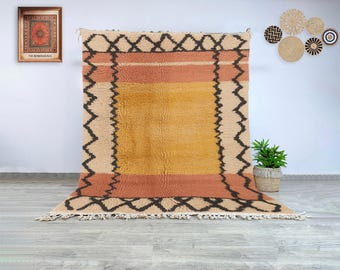 May include: A large, rectangular rug with a geometric design. The rug features a central yellow field, bordered by a peach stripe and a beige border with black zigzag patterns. The rug has a fringed edge and is displayed against a white wall.