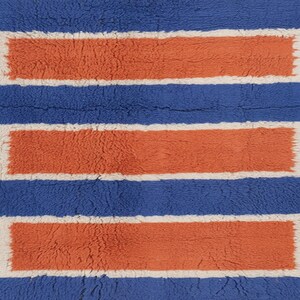 Puede incluir: Una alfombra azul con tres rect&aacute;ngulos horizontales naranjas con bordes blancos. La alfombra tiene una apariencia texturizada y afelpada, lo que sugiere una sensaci&oacute;n suave y c&oacute;moda. El dise&ntilde;o es simple pero llamativo.