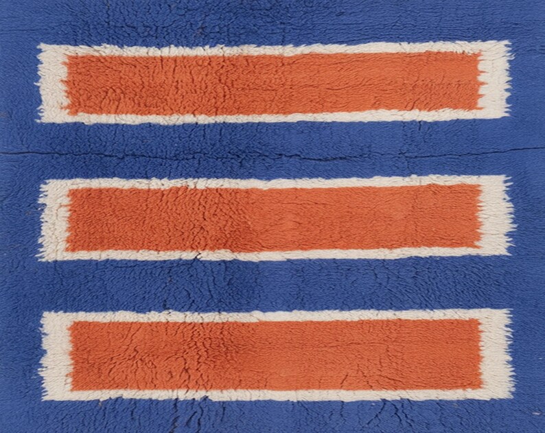 Puede incluir: Una alfombra azul con tres rect&aacute;ngulos horizontales naranjas con bordes blancos. La alfombra tiene una apariencia texturizada y afelpada, lo que sugiere una sensaci&oacute;n suave y c&oacute;moda. El dise&ntilde;o es simple pero llamativo.