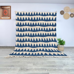 May include: A large, rectangular rug with a repeating pattern of navy blue zigzags against a cream-colored background. The rug has a fringed edge and is displayed against a white wall. The rug is a decorative home decor item.