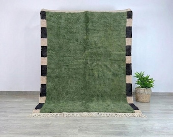 May include: A green rug with a shaggy texture, bordered by black and beige stripes on the sides. The rug has a fringe along the bottom edge. A potted plant sits to the right of the rug.