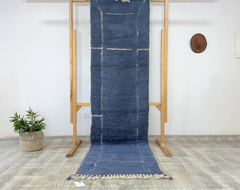May include: A long, rectangular, blue rug with a textured surface and a cream-colored geometric design. The rug is displayed on a wooden rack, showcasing its length and design. The rug has a fringe on the bottom edge.