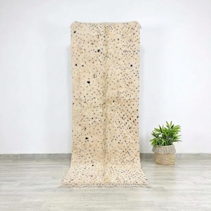 Puede incluir: Una alfombra rectangular larga con un fondo color crema y manchas negras dispersas. La alfombra tiene una textura peluda y un borde con flecos. Una pequeña planta en maceta se encuentra a la derecha de la alfombra.