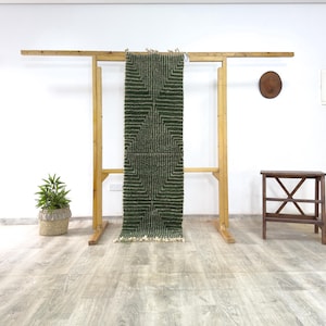 Peut inclure: Un tapis vert et beige à motifs géométriques est exposé sur un portant en bois. Le tapis a une forme rectangulaire avec un motif carré répétitif. Le tapis est fait d'une matière texturée et a des franges en bas.