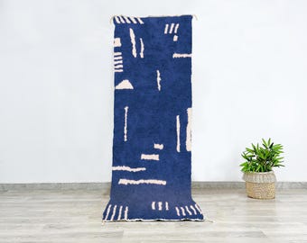 May include: A navy blue rectangular rug with a geometric pattern of white lines and shapes. The rug is displayed vertically against a white wall, with a small plant in a woven basket to the right. The rug's dimensions are not visible.