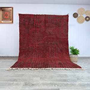 Puede incluir: Una alfombra marroquí roja y marrón con un patrón en zigzag. La alfombra tiene un borde de flecos y está colocada sobre un suelo gris claro.