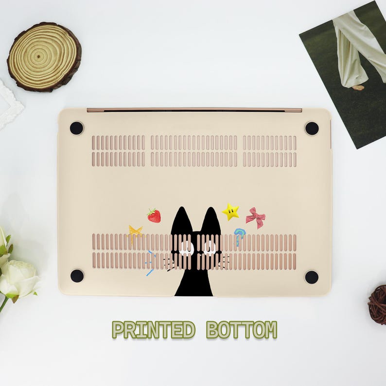 May include: A beige laptop case with a black cat peeking out from behind a fence. The case has a printed bottom with a design of a cat, strawberries, stars, and a bow.