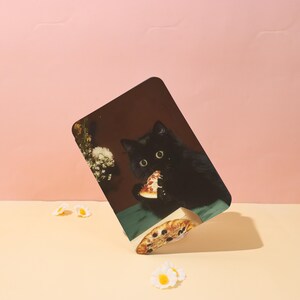 May include: A rectangular print featuring a black cat eating pizza. The cat is set against a dark brown background, with a slice of pizza in its mouth. The print is propped up on a light yellow surface with small white and yellow flower decorations.
