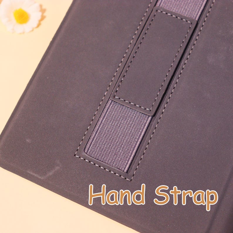 May include: A dark gray tablet case with a hand strap. The case features a stitched border and a gray elastic hand strap for secure handling. The words "Hand Strap" are printed in a light brown color on the bottom of the case.