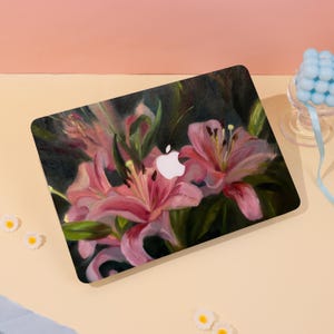 May include: Laptop cover featuring a floral painting of pink lilies with green leaves against a dark background. The Apple logo is visible. The cover has a glossy finish and is likely made of hard plastic.