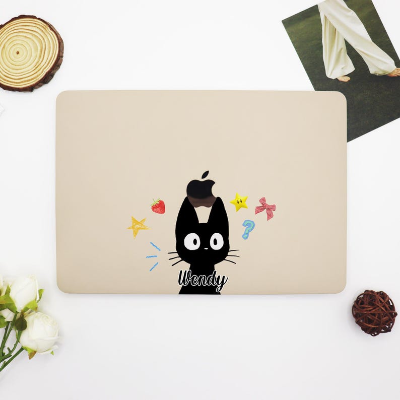 May include: A beige laptop skin with a black cat illustration and the name "Wendy" printed on it. The cat has an apple on its head and is surrounded by colorful stars, a strawberry, and a question mark.