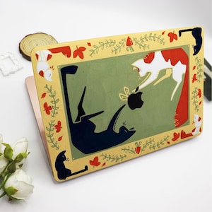 May include: A laptop skin with a yellow background and a green border. The design features two cats, one black and one white with red markings, playing with a toy. The border is decorated with red berries, green leaves, and white flowers.