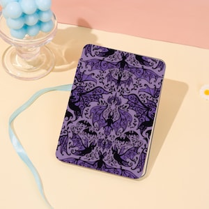 Op de afbeelding: Een rechthoekig paars notitieboek met een herhalend patroon van zwarte vleermuizen en sierlijke krullen. Het notitieboek heeft een witte rand en staat tegen een lichtroze en gele achtergrond. Een glas met blauwe bollen staat op de achtergrond.