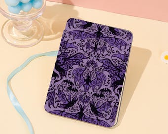 May include: A rectangular purple notebook with a repeating pattern of black bats and ornate swirls. The notebook has a white border and is set against a light pink and yellow background. A glass with blue spheres is in the background.