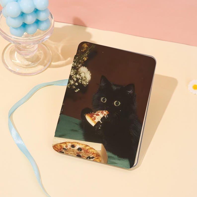 May include: A rectangular phone case featuring a black cat eating pizza. The cat has wide eyes and is holding a slice of pizza with cheese and pepperoni. A pizza box with a slice of pizza is in the foreground. The background is a dark brown.