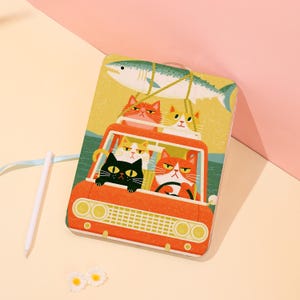 May include: A rectangular notebook featuring a colorful illustration of four cats riding in an orange car with a large fish strapped to the roof. The notebook has a light blue ribbon bookmark and is accompanied by a white pen and two small white flowers.