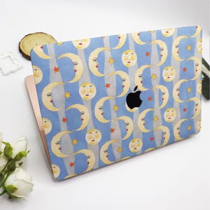 May include: A laptop with a blue cover featuring a whimsical pattern of yellow crescent moons with faces, stars, and vertical gray stripes. The Apple logo is visible. The laptop is on a white surface.
