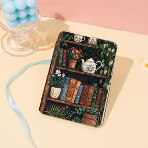 Könnte beinhalten: Eine dekorative Hülle mit einem Bücherregal-Design, das Bücher, Pflanzen und eine Teekanne zeigt. Die Hülle hat einen dunklen Hintergrund mit einem Bücherregal aus Holz, grünen Blättern und verschiedenen Topfpflanzen. Die Bücher haben verschiedene Farben und Muster.