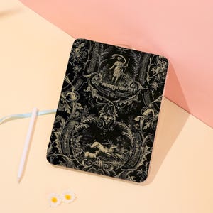 May include: A rectangular notebook featuring a black cover with a white toile-style print depicting a figure holding a spear, ornate scrollwork, and a deer in a forest scene. A white stylus and two small white flower-shaped embellishments are present.