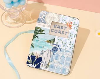 May include: A rectangular photo frame with a coastal theme. The frame features a collage of beach scenes, seashells, and floral patterns in shades of blue and white. The text "EAST COAST" is displayed in the center. A blue ribbon and a glass candle holder with blue spheres are in the background.