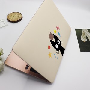 May include: A beige laptop skin with a black cat design. The cat is looking up at a falling apple and other colorful objects, including stars, a strawberry, and a key.