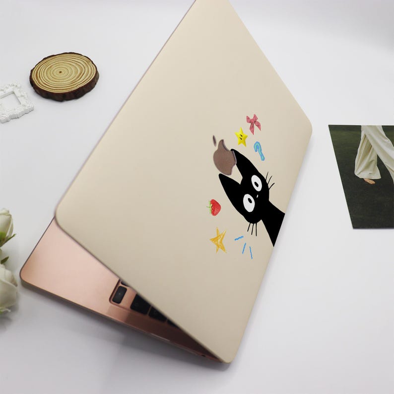 May include: A beige laptop skin with a black cat design. The cat is looking up at a falling apple and other colorful objects, including stars, a strawberry, and a key.
