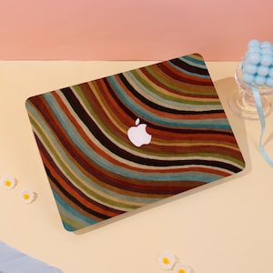 May include: A laptop with a colorful, striped design. The cover features a pattern of curved lines in shades of brown, orange, green, and blue. A white Apple logo is visible on the cover. The laptop is on a light yellow surface.