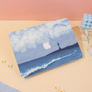May include: A laptop case featuring a coastal scene with a lighthouse, ocean waves, and a cloudy sky in shades of blue and white. The Apple logo is visible. The case is on a light-colored surface.