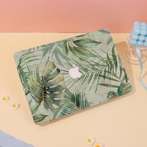 May include: A laptop with a green and gold tropical leaf design. The case features a white Apple logo and a repeating pattern of palm fronds and monstera leaves. The background is a light peach color.