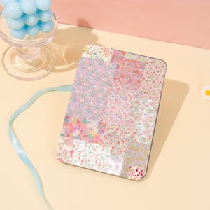May include: A rectangular, floral-patterned case with a patchwork design in shades of pink, purple, and white. A light blue ribbon and a glass candle holder with blue spheres are also visible. The case has a rounded edge.