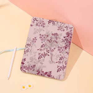 Könnte beinhalten: Eine rechteckige Tablethülle in hellem Rosa mit einem dunkelmagentafarbenen Blumenmuster aus Rosen und kleinen Blüten. Die Hülle steht aufrecht und wird von einem weißen Eingabestift sowie zwei kleinen weißen blumenartigen Verzierungen begleitet.