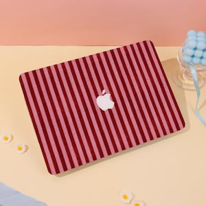 Może przedstawiać: Etui na laptopa z pionowym wzorem w paski w odcieniach czerwieni i różu. Widoczne jest białe logo Apple. Prostokątne etui zakrywa górną część laptopa.