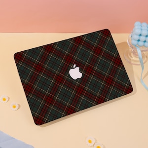 Può includere: Un laptop con un motivo a quadri verdi, rossi e oro. Il logo Apple è bianco. Il laptop è su una superficie giallo chiaro, con piccole decorazioni floreali bianche.