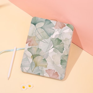 May include: A rectangular case with a watercolor-style ginkgo leaf pattern in shades of green, peach, and white. A white stylus and two small daisy-shaped decorations are next to the case on a yellow surface, with a pink background.