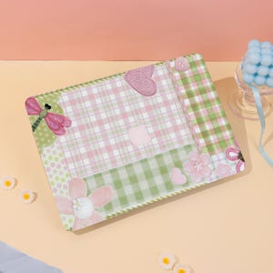 May include: A laptop skin featuring a pink and white plaid pattern combined with green and white checkered sections. The design includes decorative elements such as a pink dragonfly, various pink flower shapes, small heart icons, and a button.