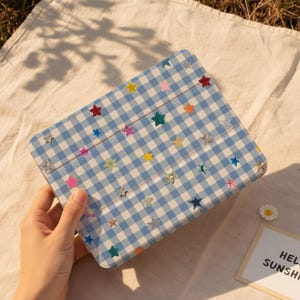 May include: A blue and white gingham tablet case adorned with colorful star-shaped stickers. The case is held in a hand and is open, revealing the interior. The overall aesthetic is cheerful and playful.
