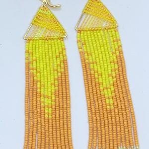 May include: A pair of long dangle earrings featuring a gold-tone triangular frame wrapped in yellow thread. Below the frame, multiple strands of small, vibrant yellow and orange seed beads hang down, creating a chevron pattern in the beadwork.