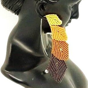 May include: A long dangle earring featuring four stacked leaf-shaped macrame elements in a gradient of colours, transitioning from light yellow at the top to orange, rust, and dark brown at the bottom, suspended from a thin silver-toned metal wire.