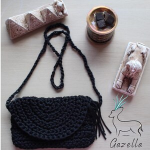 May include: A black crochet crossbody bag with a flap and fringe. The bag has a long, braided strap. The image also shows decorative items, including a pyramid-shaped sculpture and a small bowl.